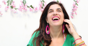 Luciana Peker se retrata en una foto. Es una mujer de cabello cafe obscuro, y largo, más abajo de sus hombros.  Está sonriendo con vista hacía al horizonte y arriba, mano levantada a la mejilla de su cara, con sonrisa grande y boca abierta. Viste una camiseta verde con aretes grandes y pulseras de muchos colores. En el fondo contra pared hay seis ramitas de flores color rosa pálida con hojas verdes, colgadas cada rama de manera deocrativa, hojas y pétalos hacía abajo.